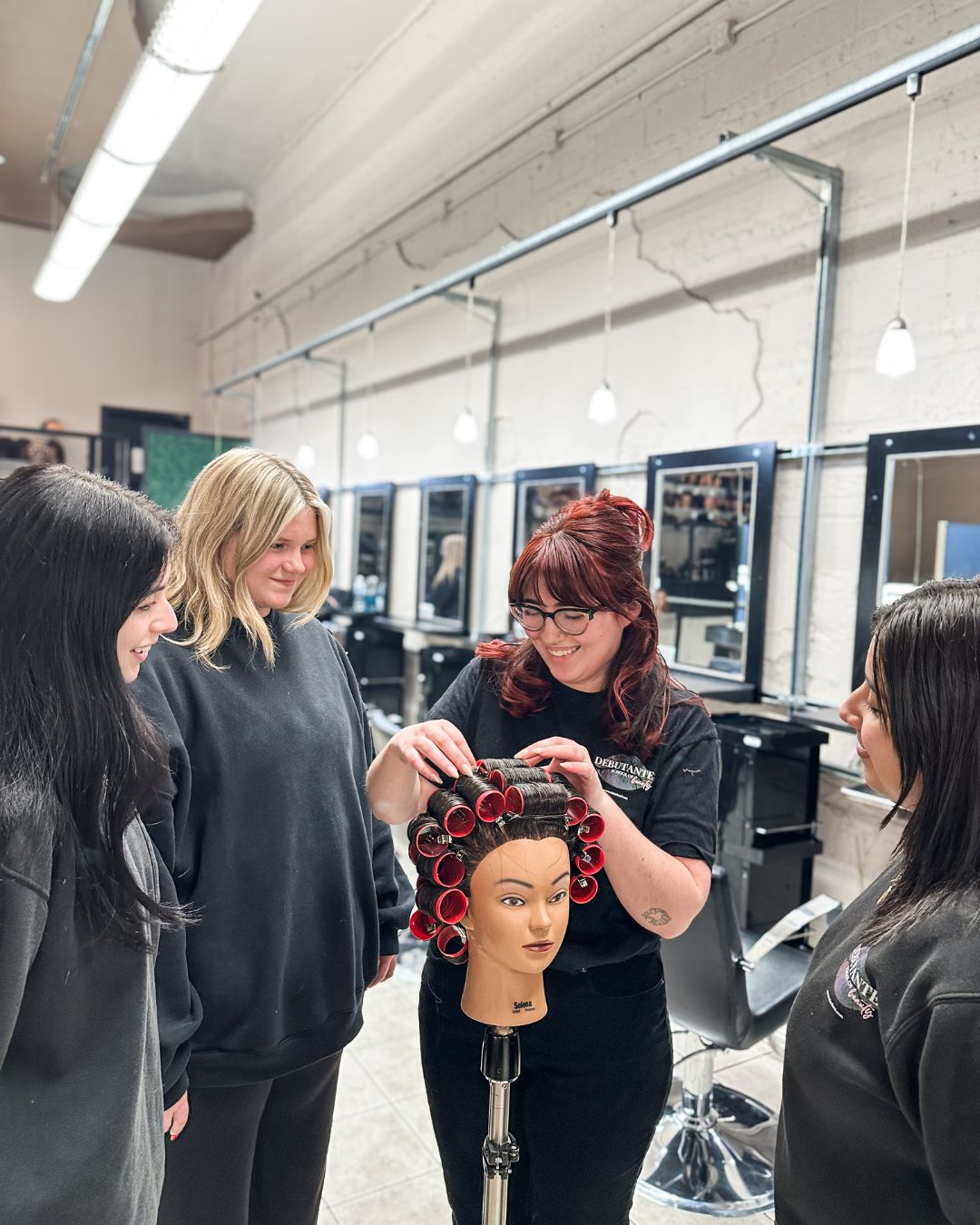 A young female with red hair teaching a roller set to a small group of students.